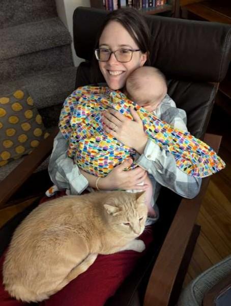 Cora smiles at the camera, sitting and holding her daughter over her shoulder with a cat on her lap.