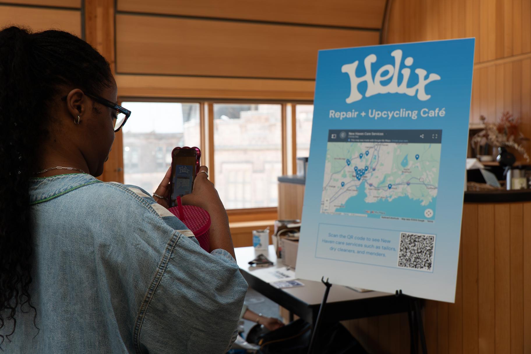 A person takes a photo of a poster that reads "Helix Repair + Upcycling Cafe" in white text on a blue background, with a map with pins showing locations.