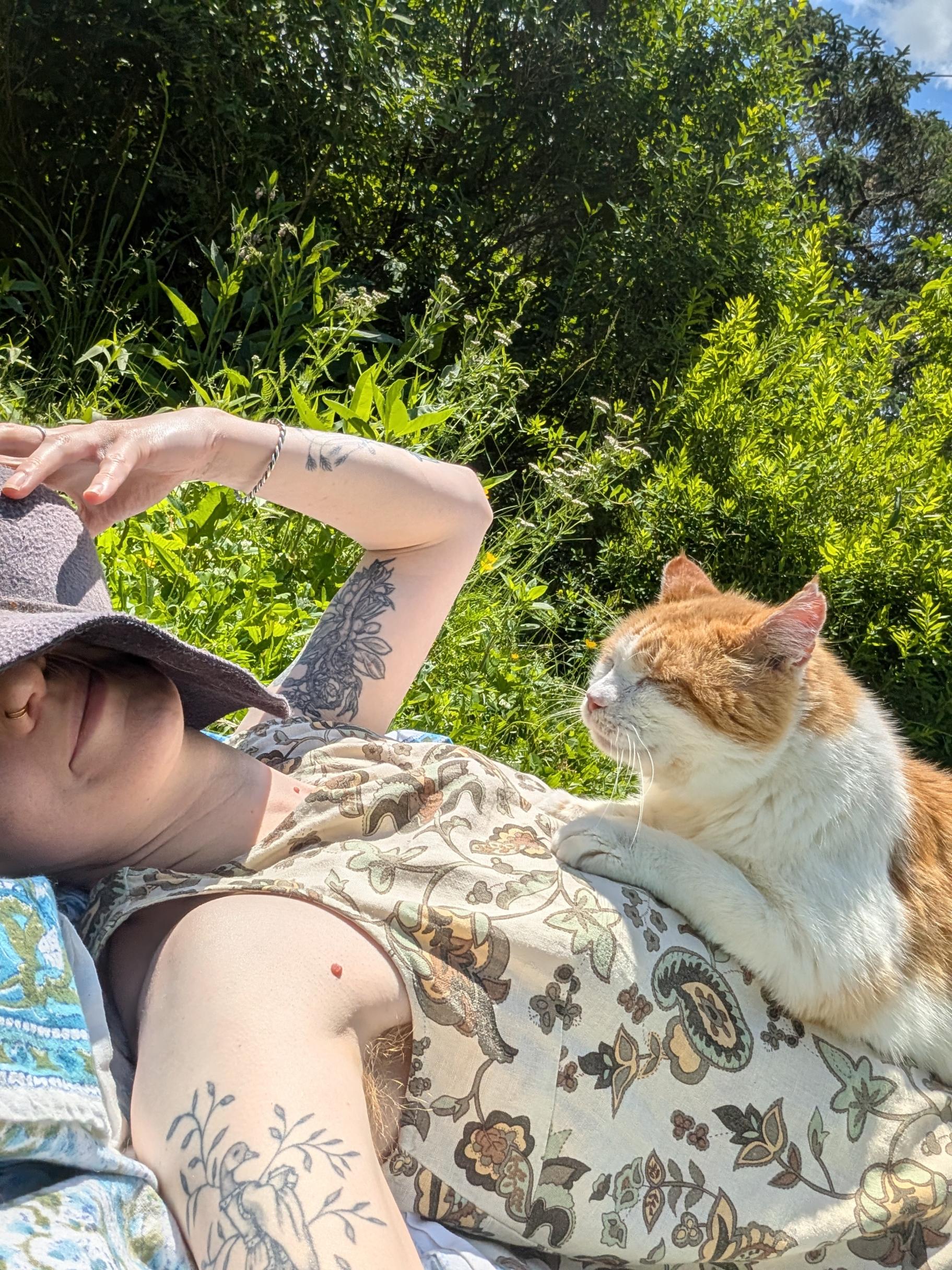 Laine is laying in the grass with a cat on their lap