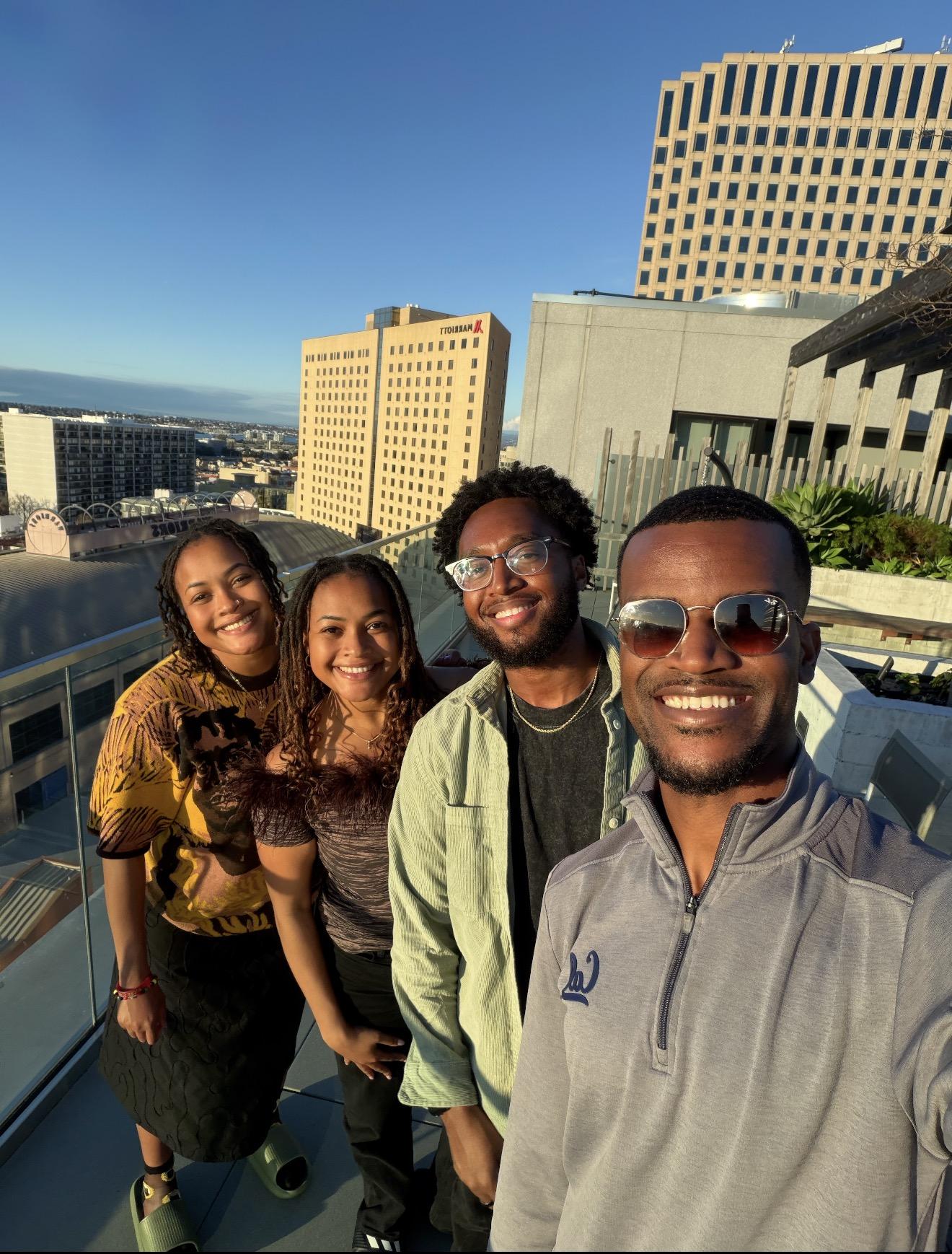 Mica and Maya Caine stand with two men, smiling for a selfie in a rooftop patio. Maya is wearing the feathered top described in the story.