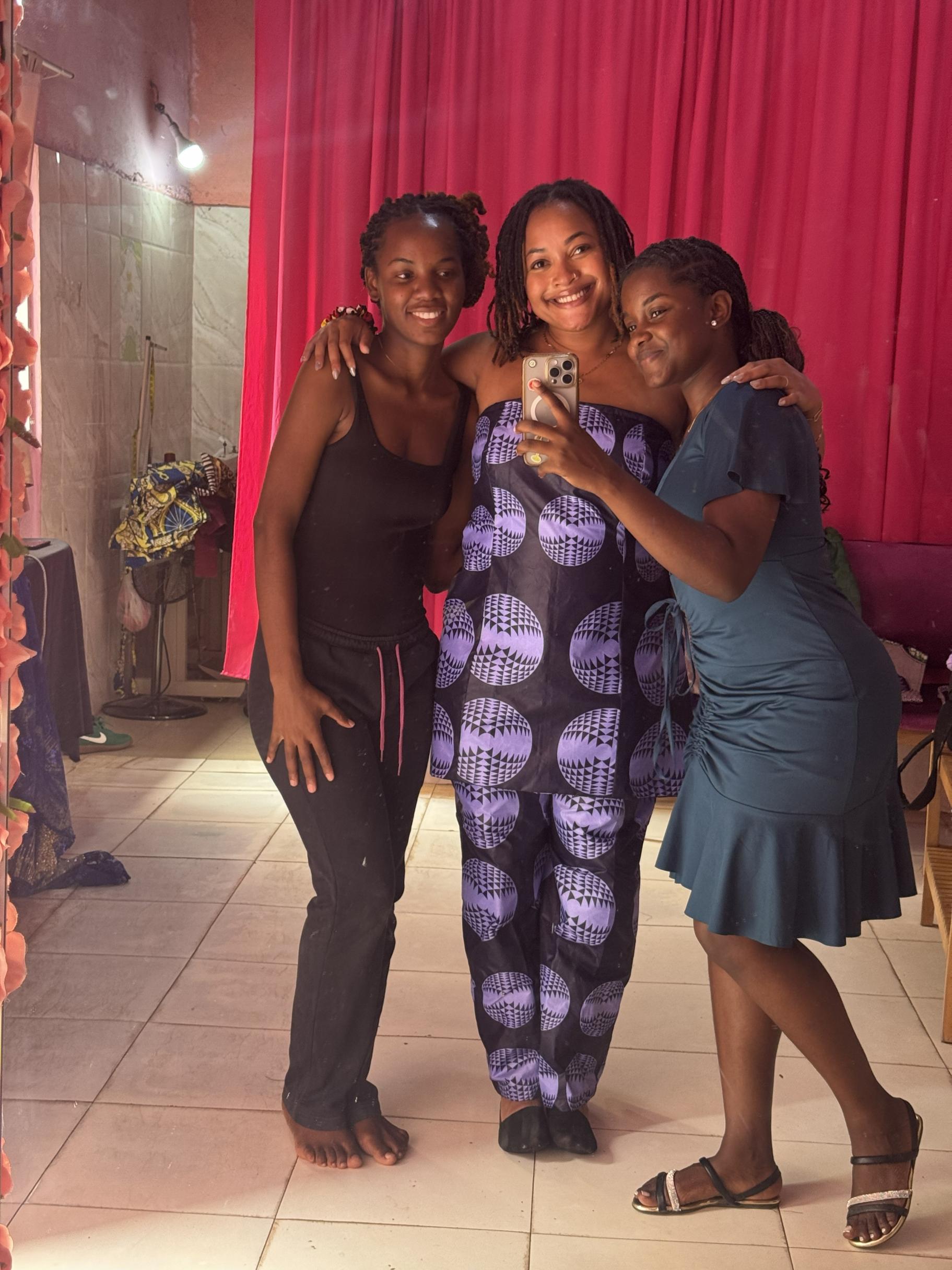 Mica is in the center of two other women for a mirror selfie, wearing a custom garment in a purple, white and black circular fabric.