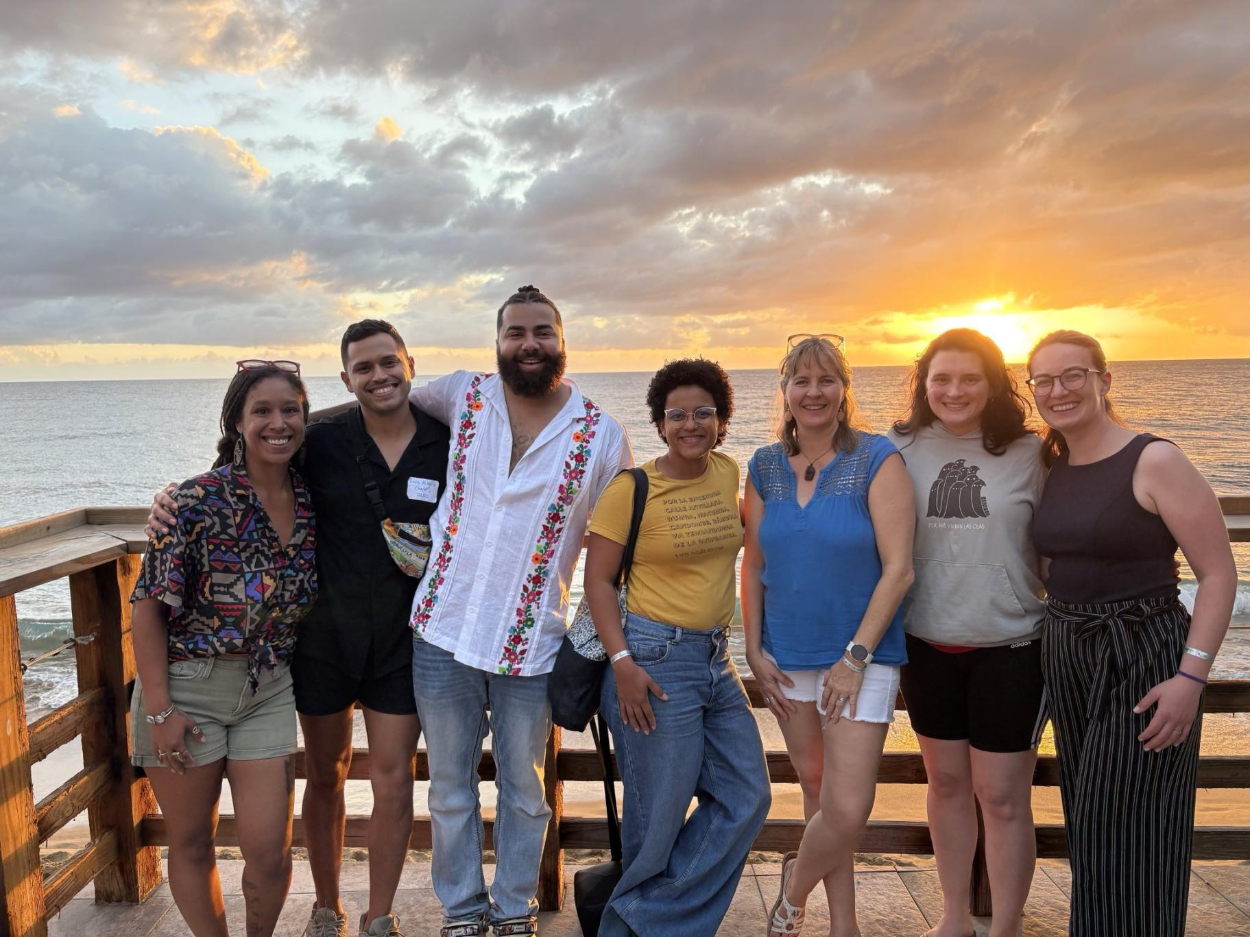 Switzer Fellows on the planning committee stand together on the deck at Villa Cofresí, with the ocean and an orange sunset in the background. From left to right: Olivia Walton, Luis Alexis Rodríguez-Cruz, AJ Hudson, Christina De Jesús Villanueva, Carina Bracer, Sara Santiago, and Chelsi Sparti.