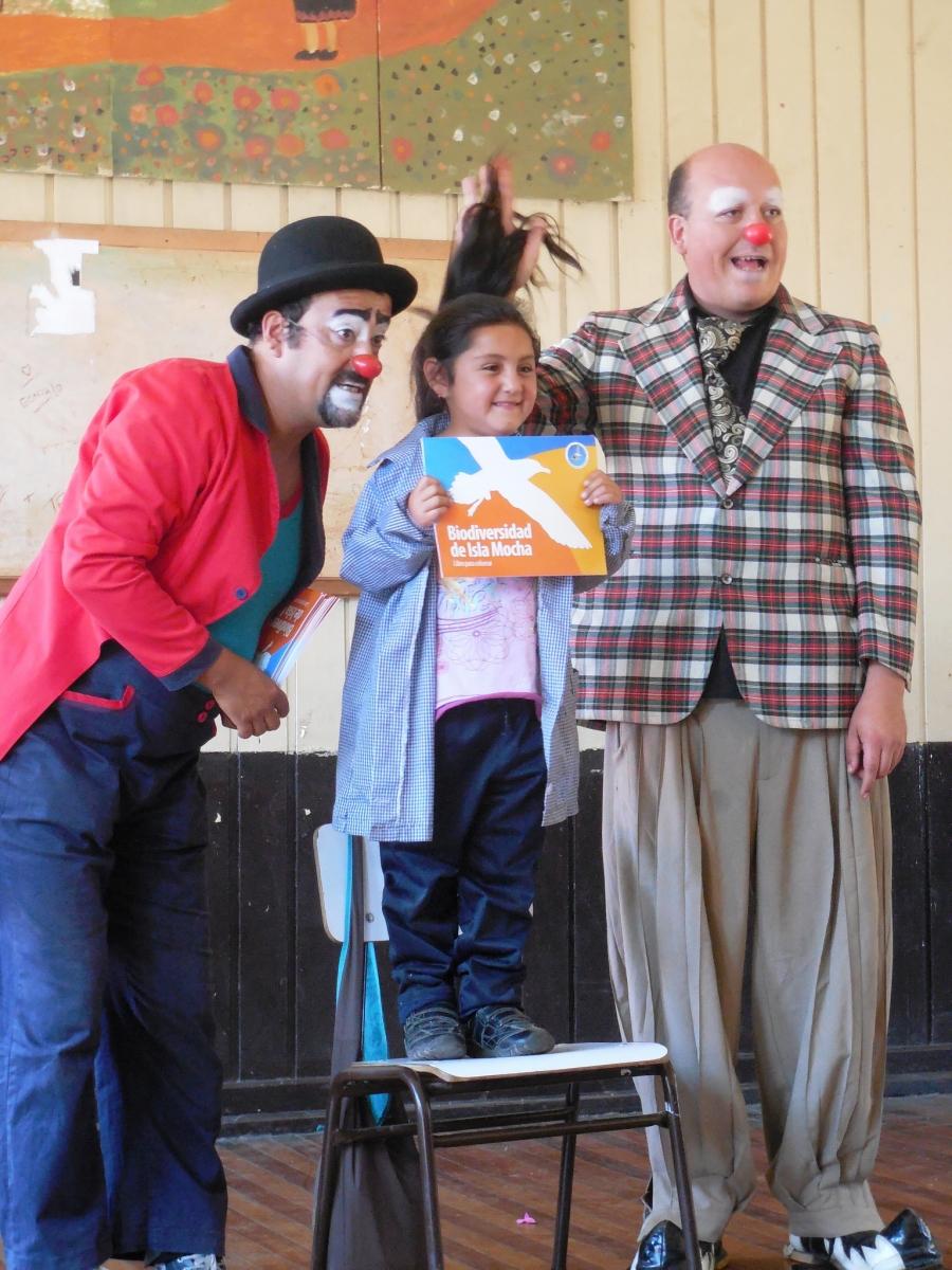 Gonzalo Diaz (left) and Miguel Muñoz (right), the Mocha clowns, awarding a coloring book about the biodiversity of Isla Mocha to a Mochan elementary schooler
