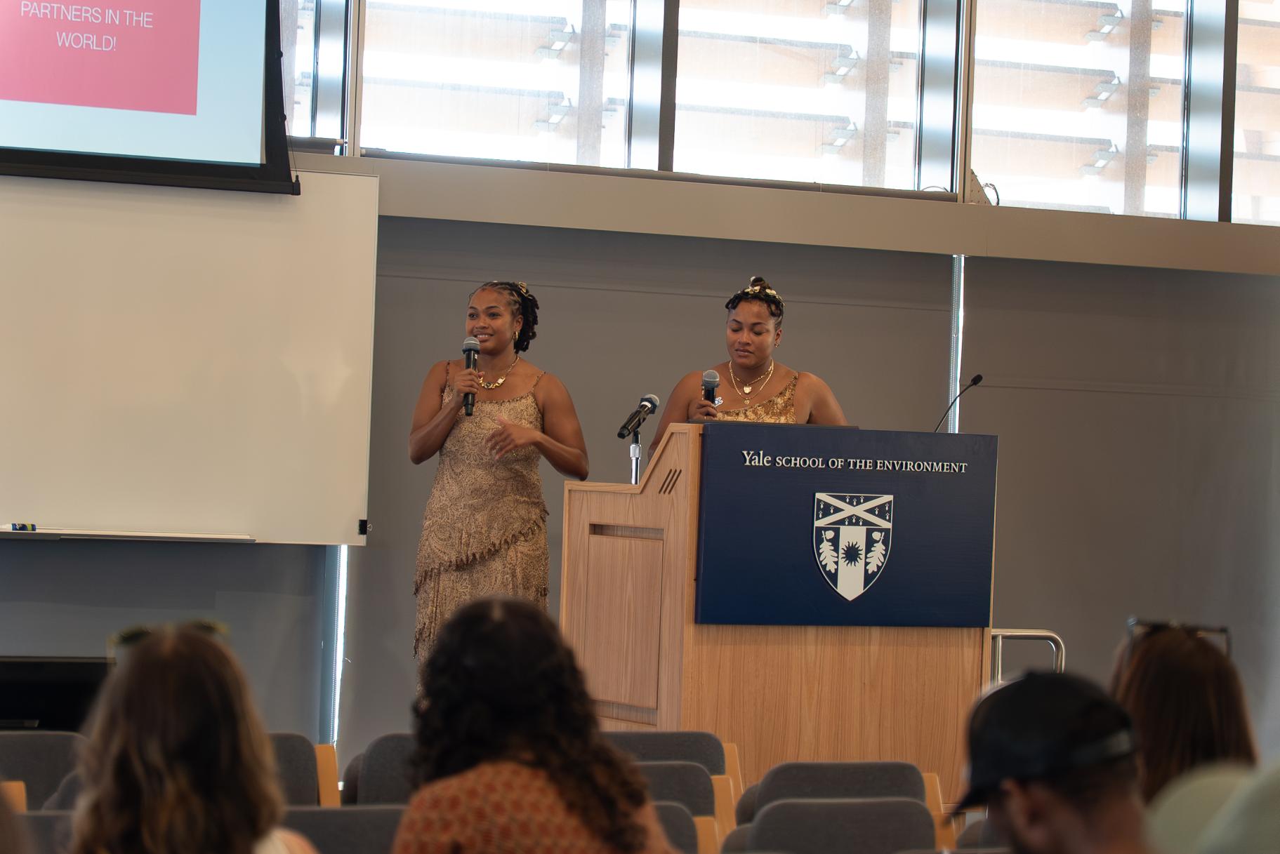 Maya and Mica stand in beautiful beige dresses at a podium with YALE SCHOOL OF THE ENVIRONMENT written on the front.