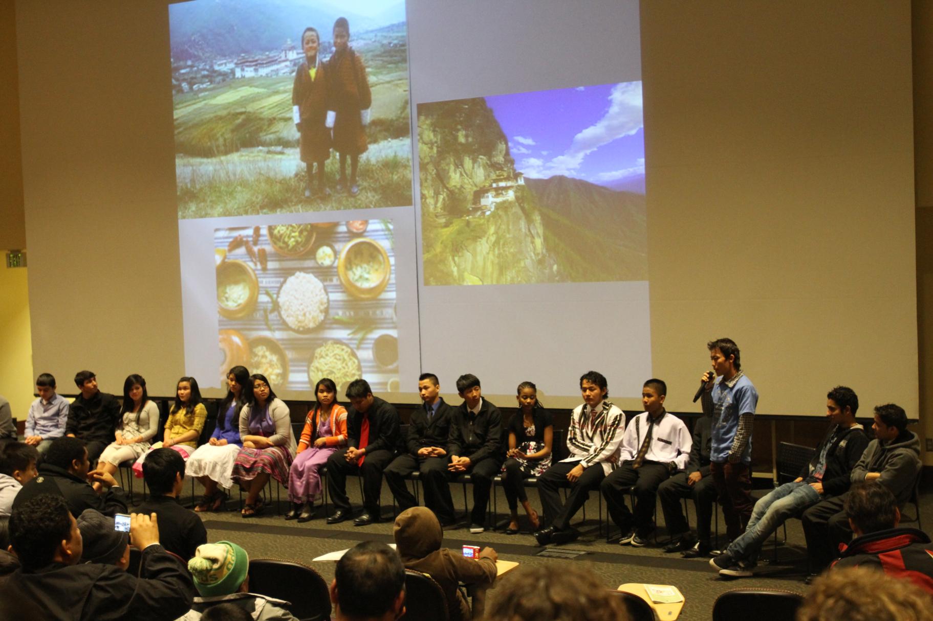 People sit in a line on a stage in front of a projector showing images of a young boy, a landscape and a bowl of food. A young man stands and speaks into a microphone.