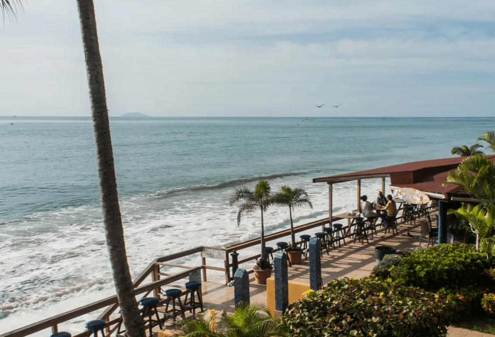 A patio with people, palm trees, plants, tables, chairs and umbrellas overlooks the ocean.